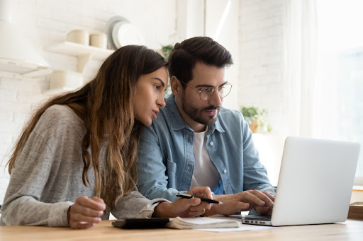 A man and woman sitting at a kitchen table with a laptop and notepad open in front of them.