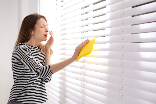 A woman cleaning window blinds with a cloth. She looks like she is about to sneeze.