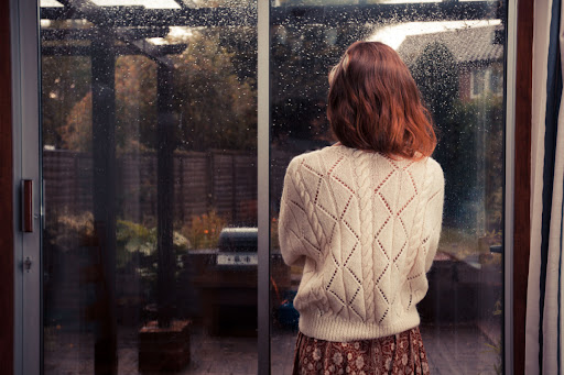 A woman standing in front of a window and looking out at the rain.