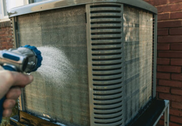 A close-up of a hand using a hose to clean an outdoor AC unit.