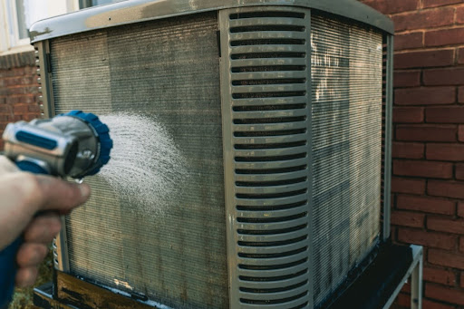 A close-up of a hand using a hose to clean an outdoor AC unit.