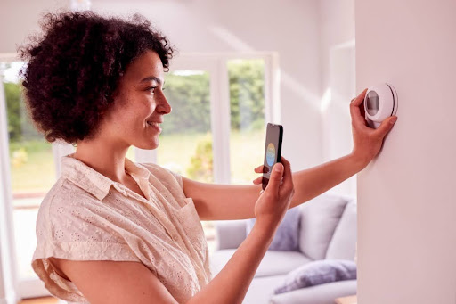 A smiling woman adjusting a smart thermostat in a home with a smartphone.